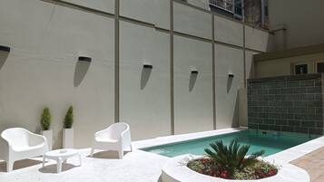 Un espace aménagé au bord de la piscine, avec une piscine blanche, des chaises blanches, une petite table et un bac à fleurs orné de fleurs rouges.