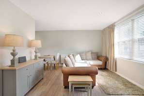 A modern living room with a sofa, a coffee table, a dining table with chairs, a lamp, and a radiator. - Cosy Keswick 1BR|Sleeps 2| by Stay with Us (Keswick)