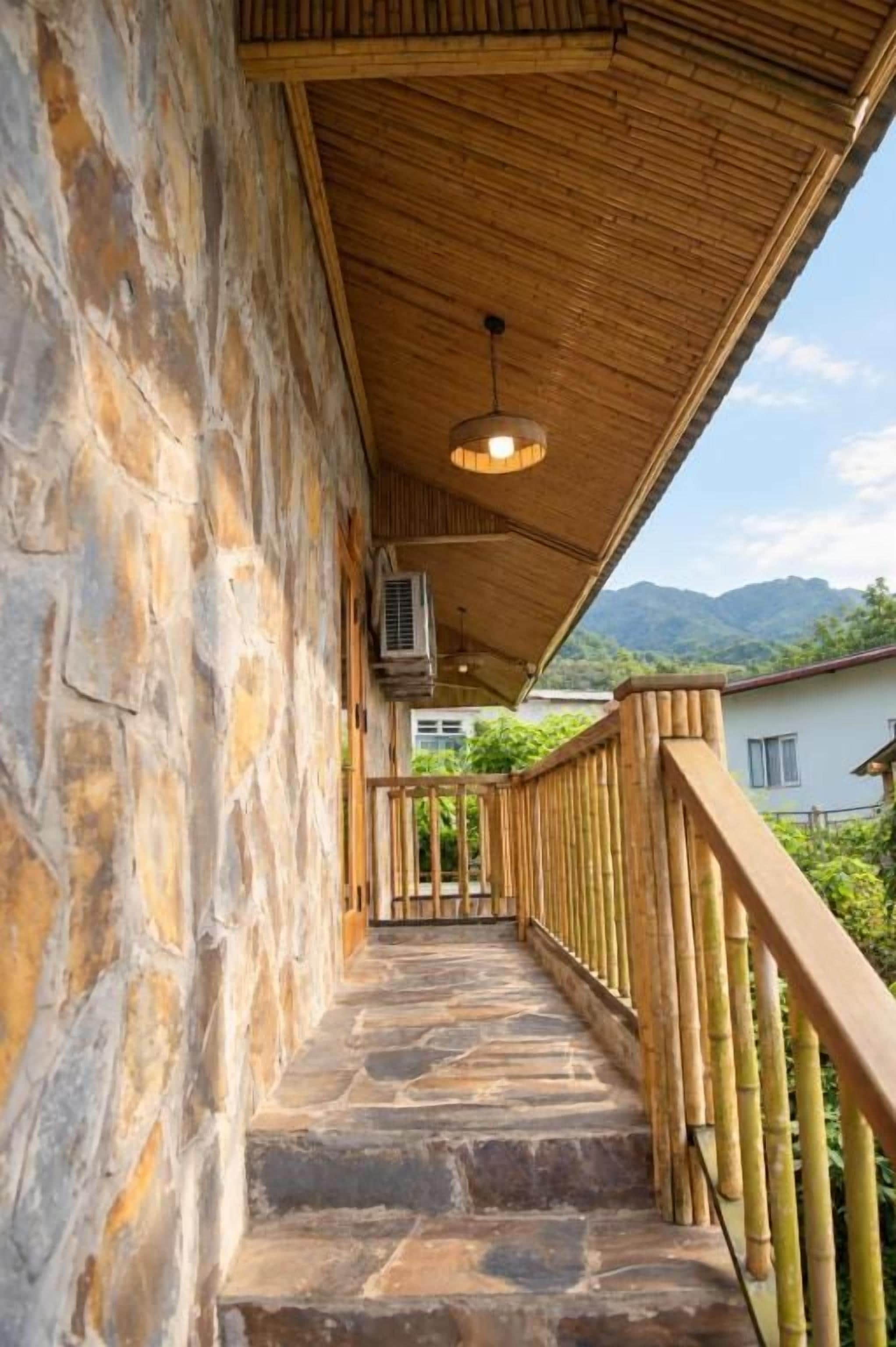Una escalera de piedra conduce a una zona cubierta con barandilla de bambú, una unidad de aire acondicionado y vistas a la vegetación y montañas a lo lejos.