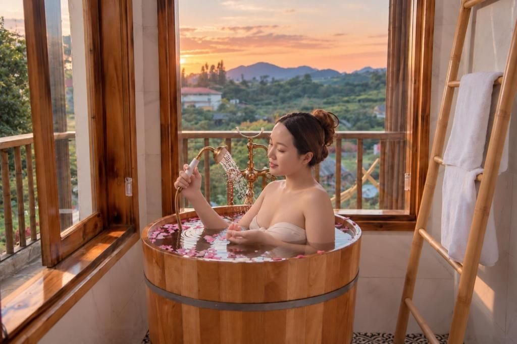 Una persona en una tina de madera llena de agua y pétalos de rosa, con vistas a montañas y follaje a través de amplios ventanales.