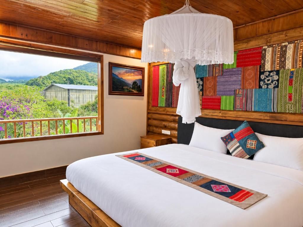 A bedroom with a large bed, a wooden headboard, a hanging white canopy, a view of greenery outside, and a wall decorated with colorful textiles.