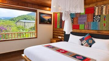 A bedroom with a large bed, a wooden headboard, a hanging white canopy, a view of greenery outside, and a wall decorated with colorful textiles.