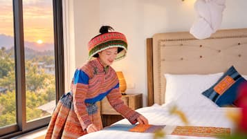 A person in traditional attire arranging a patterned textile on a bed in a room with a view of a mountainous landscape.