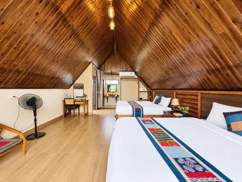 Una habitación con techo inclinado, una cama con colcha colorida, un escritorio con silla y un ventilador.