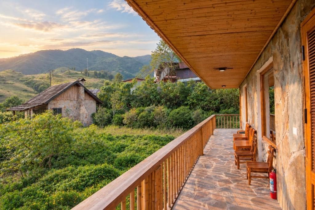 A wooden balcony with stone flooring, overlooking a lush green landscape with hills in the background.