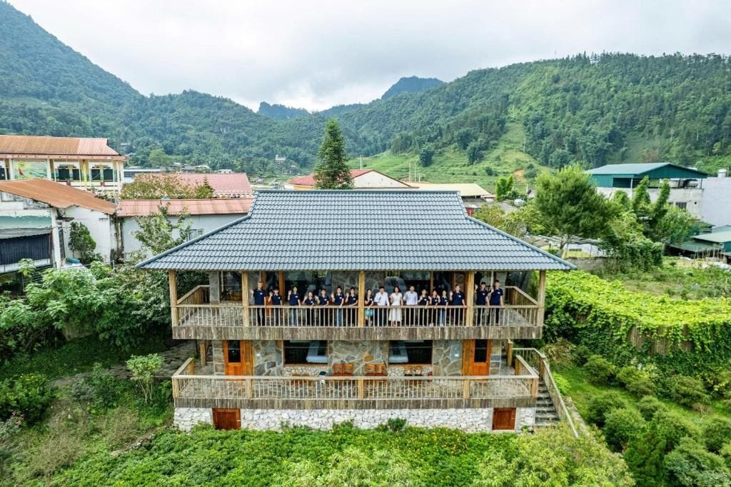 Un edificio de varios pisos con balcón, rodeado de vegetación exuberante y montañas al fondo.