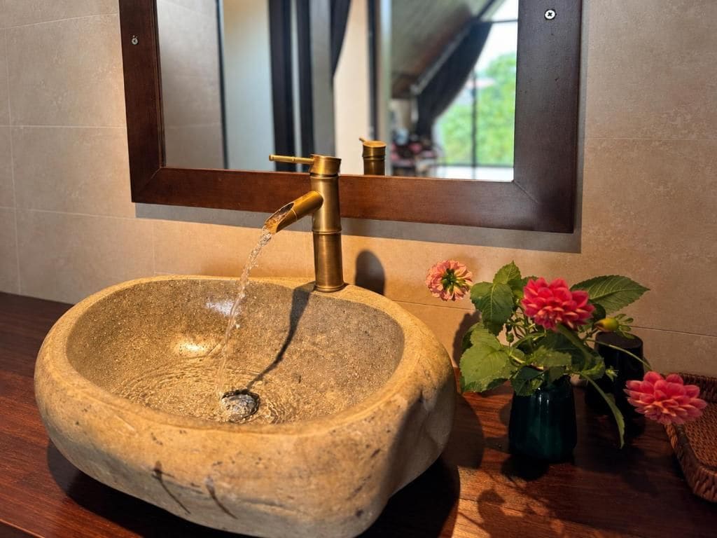 Un lavabo de piedra con un grifo de latón, un espejo encima y un florero con flores rosas en la encimera.