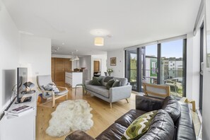 A modern living room with a sofa, a chair, a coffee table, and a television. There is a large window providing a view of the outside. - The Water of Leith Lookout (Edinburgh)