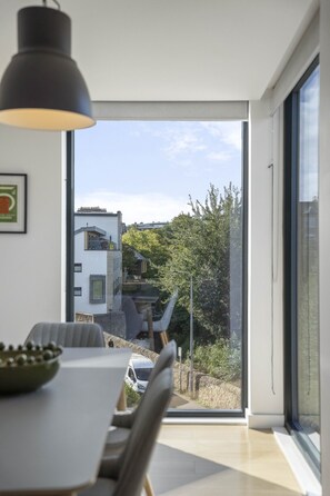 A modern dining area with a table and chairs, a pendant light, and a view of a building and greenery through a glass door. - The Water of Leith Lookout (Edinburgh)