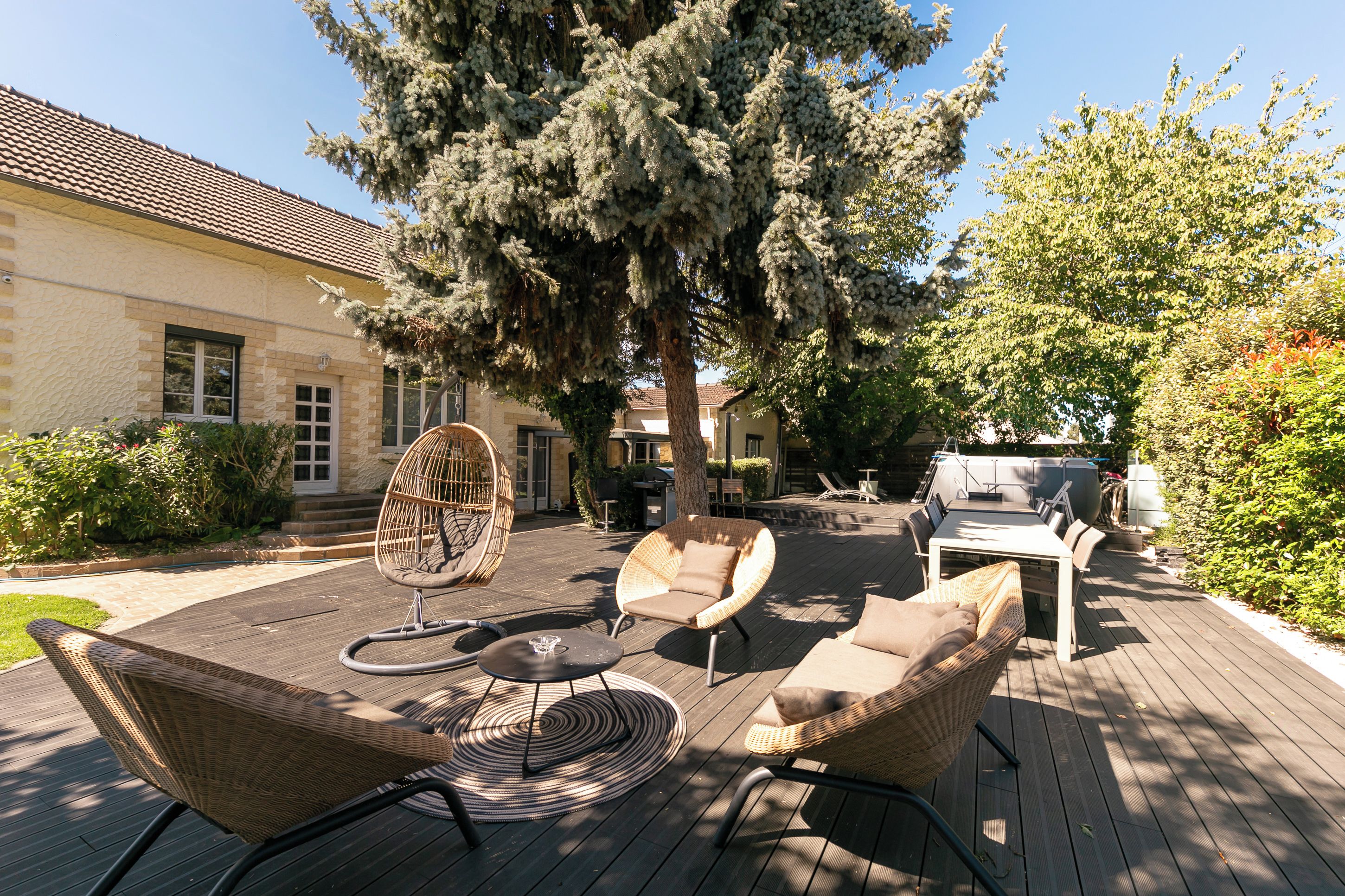 An outdoor patio with wicker furniture, a round table, and a large tree providing shade.