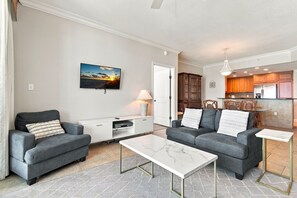 A living room with two grey sofas, a white coffee table, a flat-screen TV mounted on the wall, and a kitchen in the background. - Portofino 1-1808 🌊🏖️ Gulf Views from the 18th Floor | Spa + Tennis Onsite (Pensacola Beach)