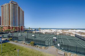 A tennis court adjacent to a beach with a high-rise hotel in the background. - Portofino 2-1101 🌊🌅 11th Floor Gulf Views | Gulf Front Pool + Sauna (Pensacola Beach)
