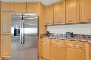 A kitchen with wooden cabinets, a stainless steel refrigerator, and a granite countertop. - Portofino 2-1101 🌊🌅 11th Floor Gulf Views | Gulf Front Pool + Sauna (Pensacola Beach)