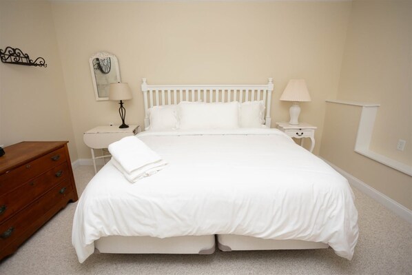 Een keurig opgemaakt bed met witte beddengoed, een houten dressoir, twee nachtkastjes met lampen, en een aan de muur hangend schilderij.