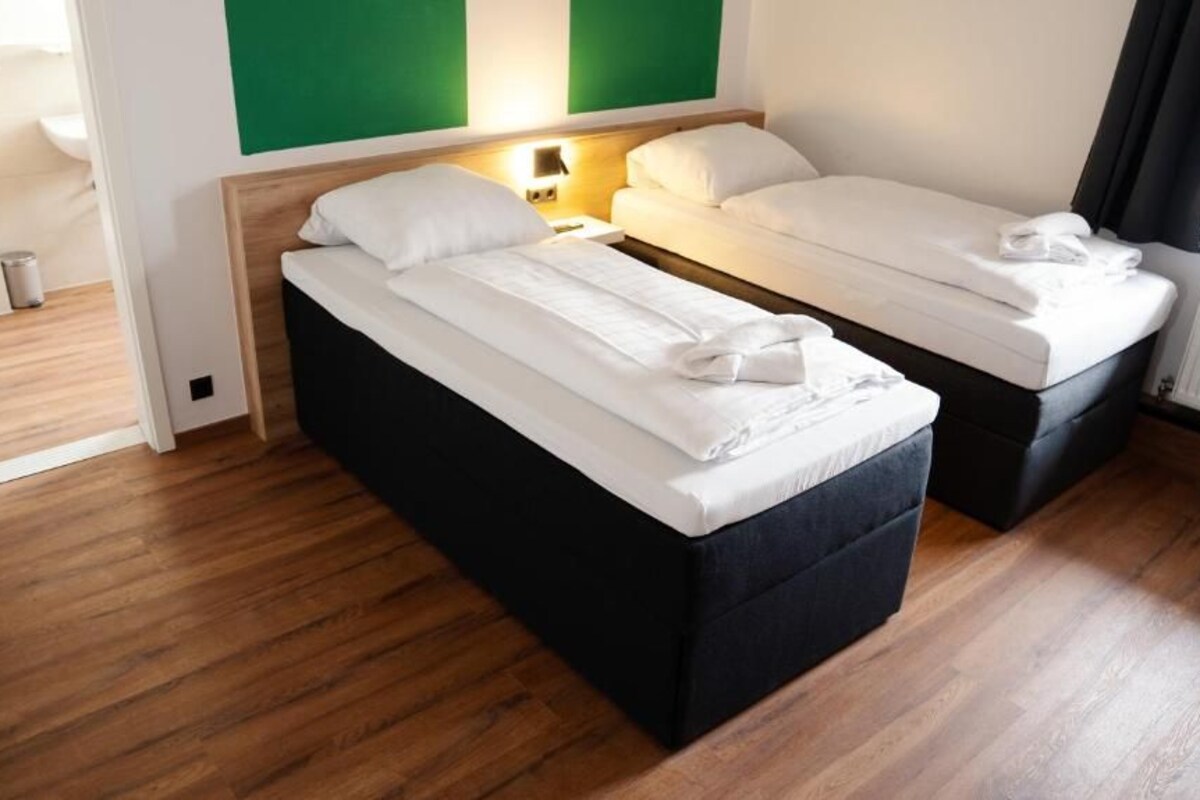 A hotel room with two beds, a wooden floor, a green accent wall, and a nightstand with a lamp.