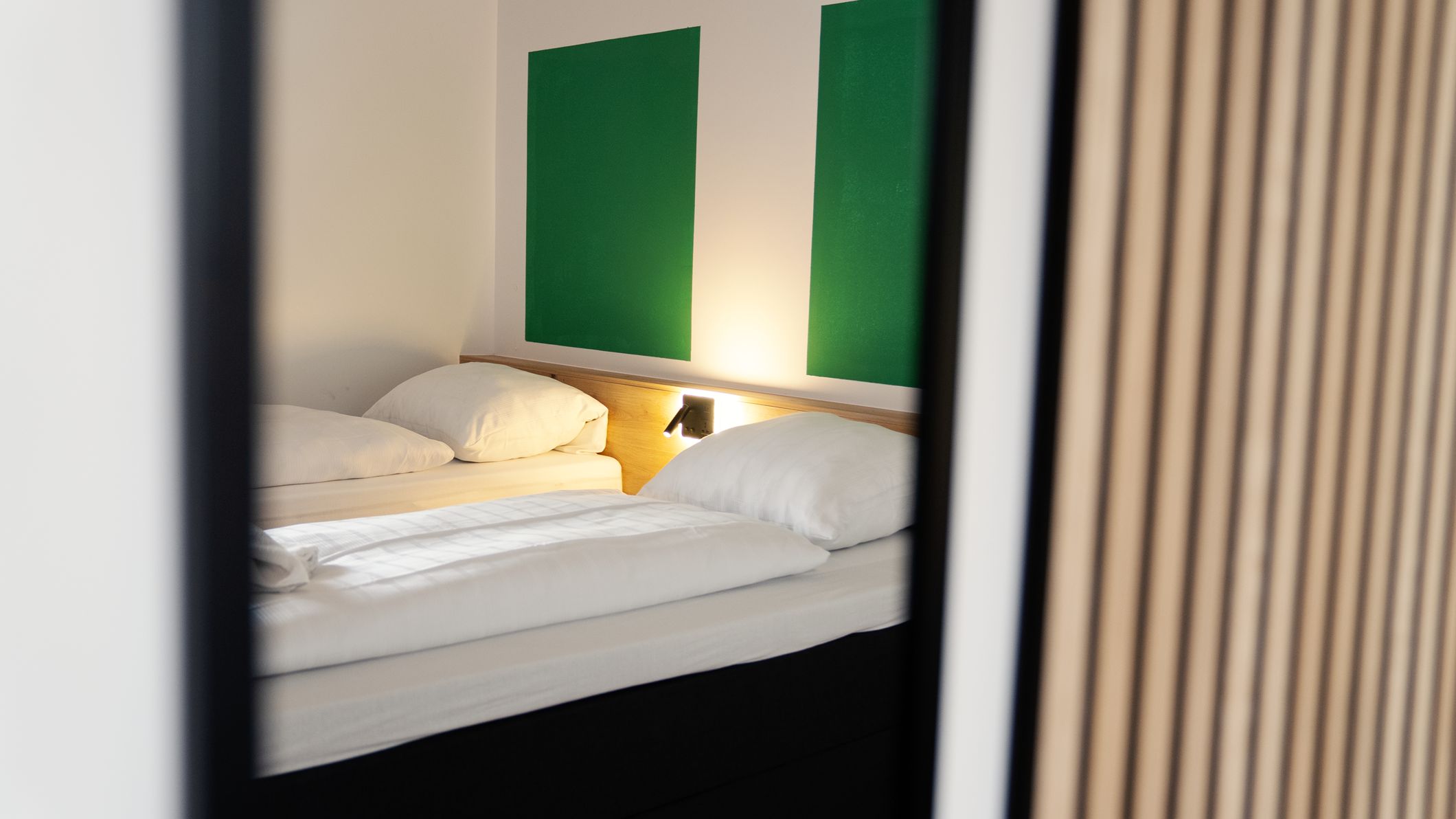 Una cama con ropa de cama blanca, una mesita de noche con lámpara y un panel de pared verde.