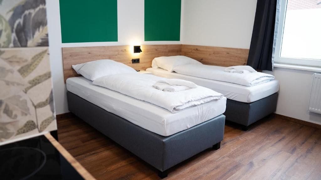 A room with two beds, a wooden headboard, a wall-mounted light, and a window with curtains.