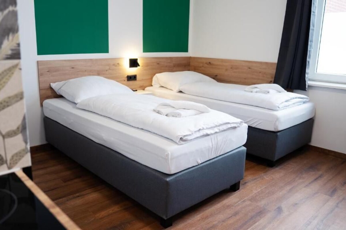 A room with two beds, a wooden headboard, a wall-mounted light, and a window with curtains.