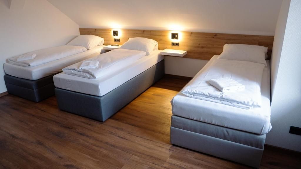 Une chambre d’hôtel avec trois lits simples, chacun doté d’une table de chevet et d’une lampe. La chambre est pourvue d’un plancher en bois et d’un plafond en pente.