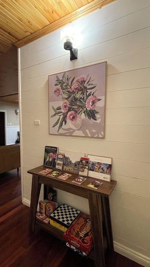 A room with a wooden ceiling, a wall-mounted light fixture, a framed floral artwork, a small table with travel brochures, and a checkered board game. - Pemby Nest (Pemberton)