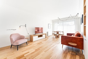Een moderne woonkamer met houten vloer, een rode bank, een roze fauteuil, een televisie op een houten dressoir, en een eethoek met tafel en stoelen.