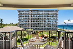 A balcony with a table set for dining, overlooking a large hotel building and a beach. - Whaler 413 Oceanview Studio Condo Apts (Lahaina)