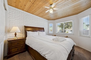 Un dormitorio con techo de madera, una cama con ropa de cama blanca, una mesita de noche con lámpara y ventanas con persianas.
