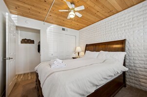 Un dormitorio con techo de madera, pared de ladrillo blanco, estructura de cama de madera y ventilador de techo.