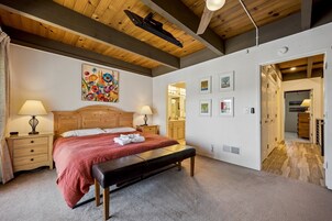 Un dormitorio con cabecera de madera, un banco, una cama con manta roja, dos mesitas de noche con lámparas y una pared con cuadros enmarcados.