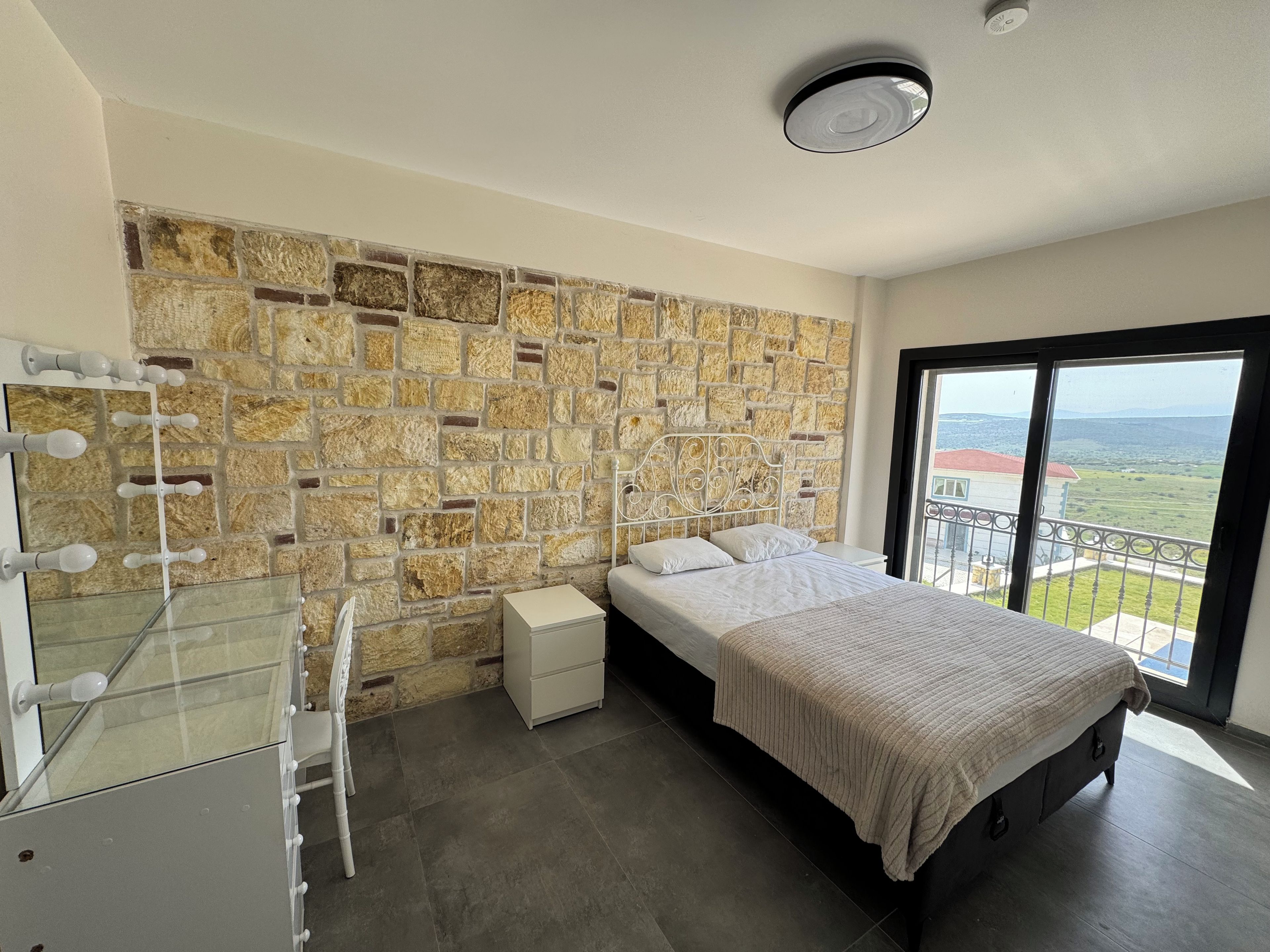 Une chambre avec un mur en pierre, un lit, une commode et une grande fenêtre donnant sur un beau paysage.