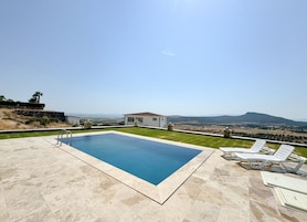 Una piscina rectangular con vistas despejadas del paisaje circundante, un pequeño edificio y algunas sillas de descanso.