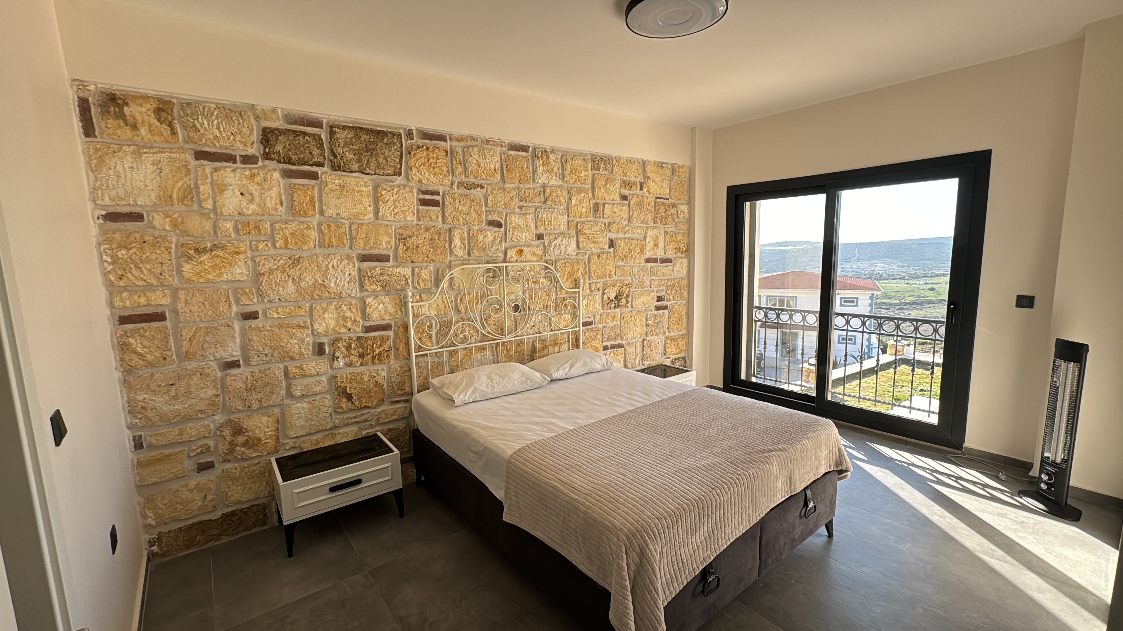 A bedroom with a stone wall, a bed with a beige blanket, a nightstand, and a large window with a view of buildings.