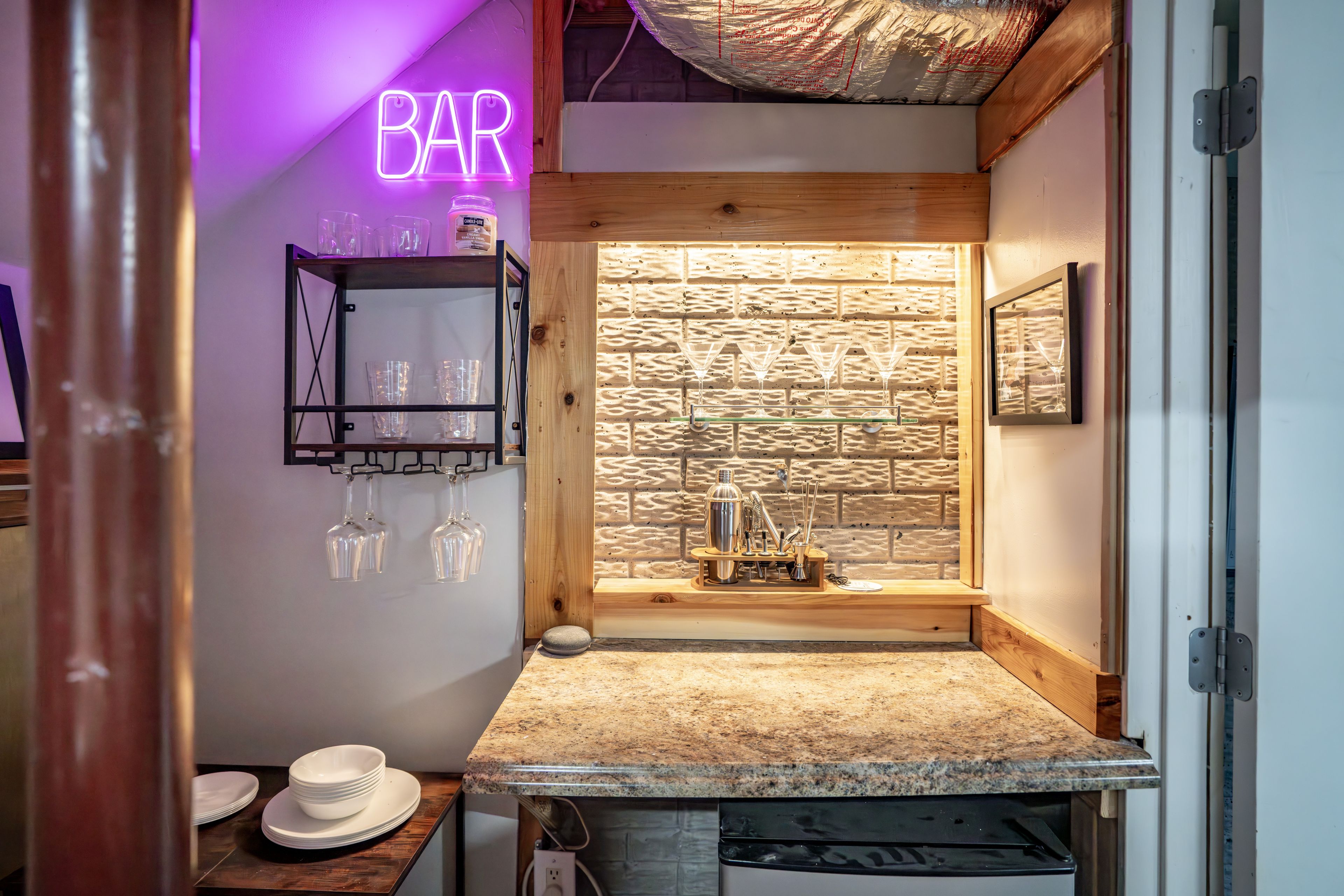 Una zona de bar con un letrero de neón, una pared de madera y una barra con vasos y un coctelero.