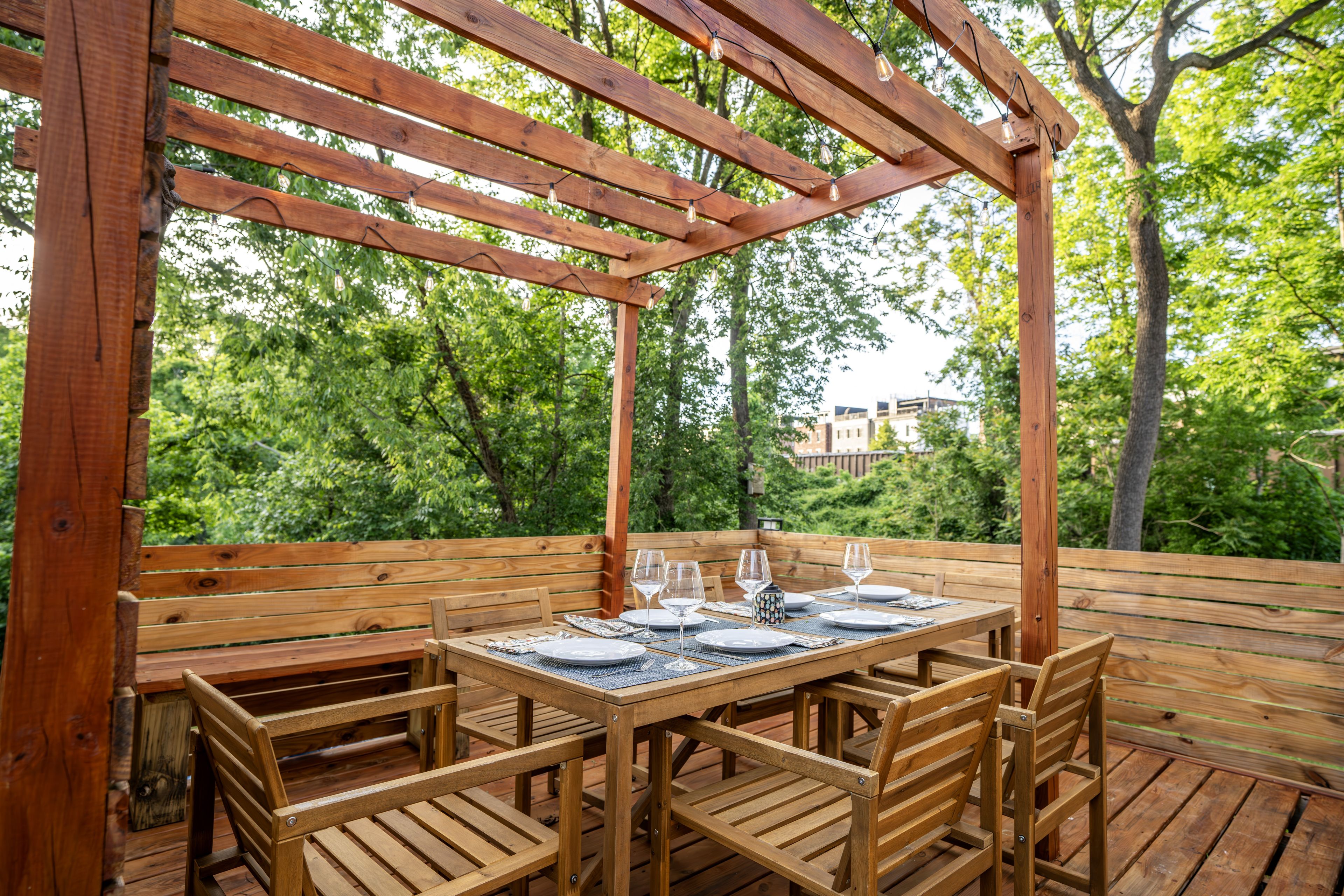 Una pérgola de madera al aire libre con una mesa dispuesta para comer, rodeada de sillas.