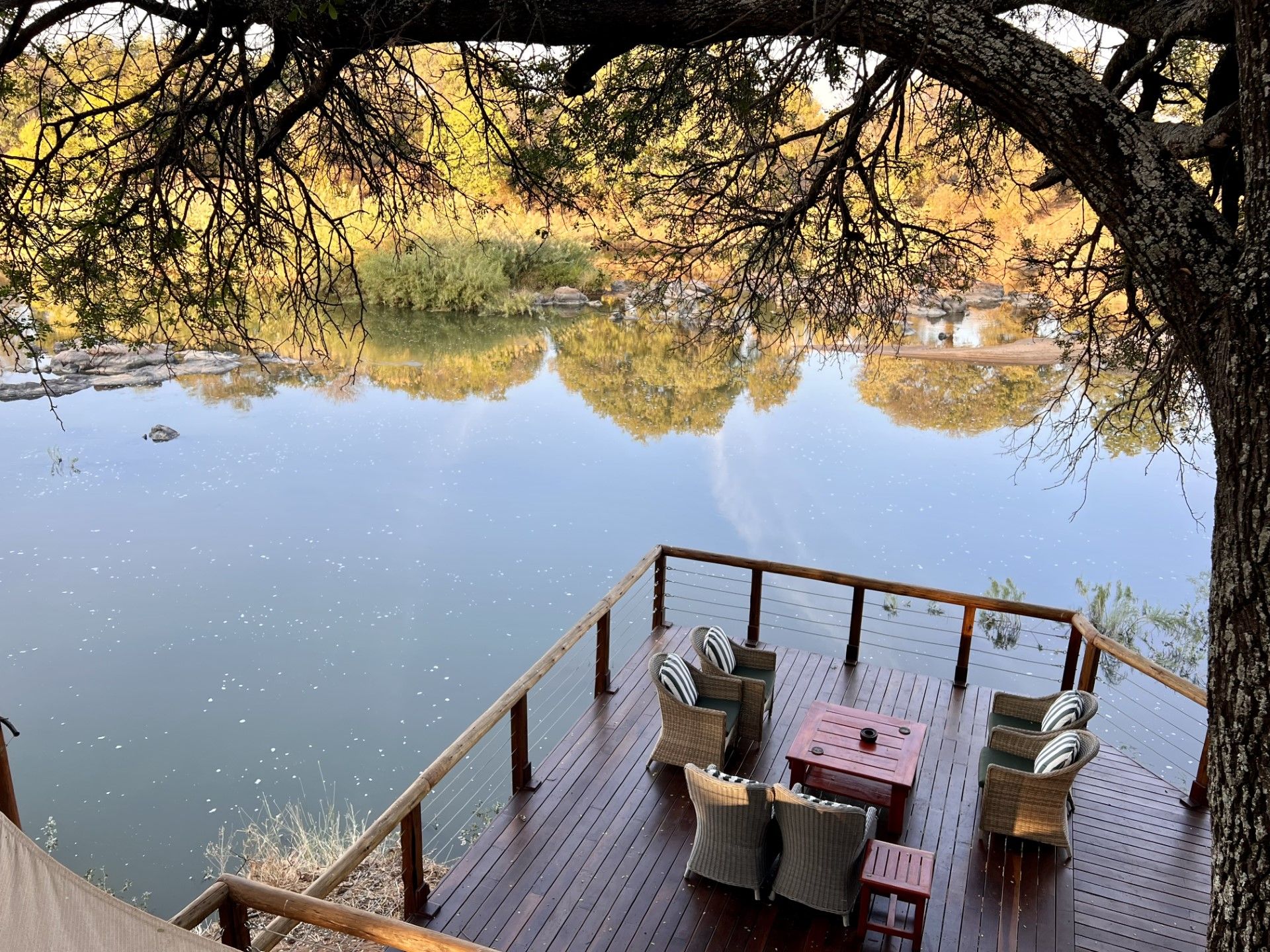 Une terrasse en bois, agréée de mobilier d’extérieur, qui offre une vue sur une étendue d’eau paisible, le tout étant entouré d’arbres.