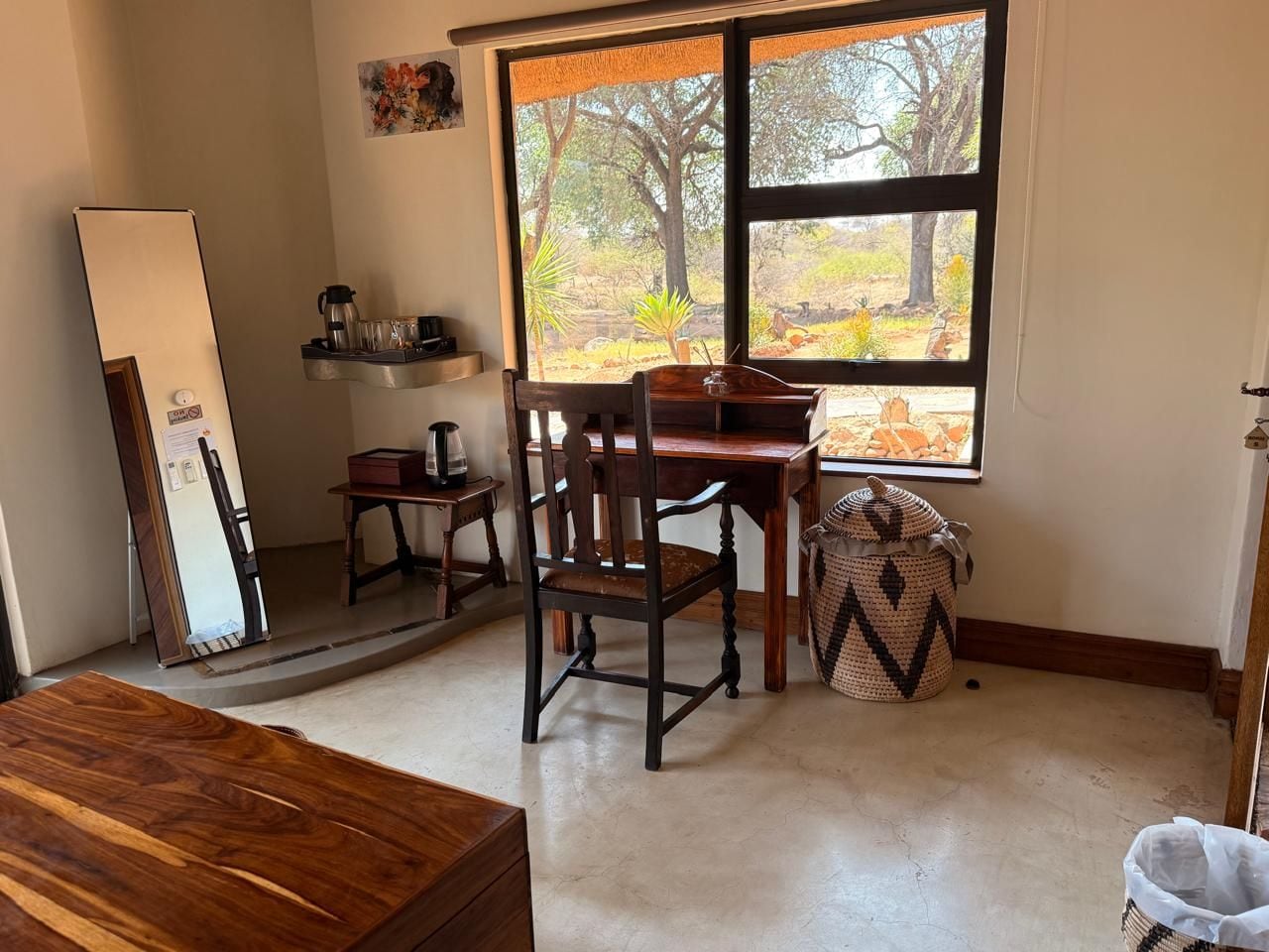 Una habitación con una mesa de comedor de madera, una silla, un estante pequeño con una cafetera, un ventanal con vistas a árboles y una cesta tejida.