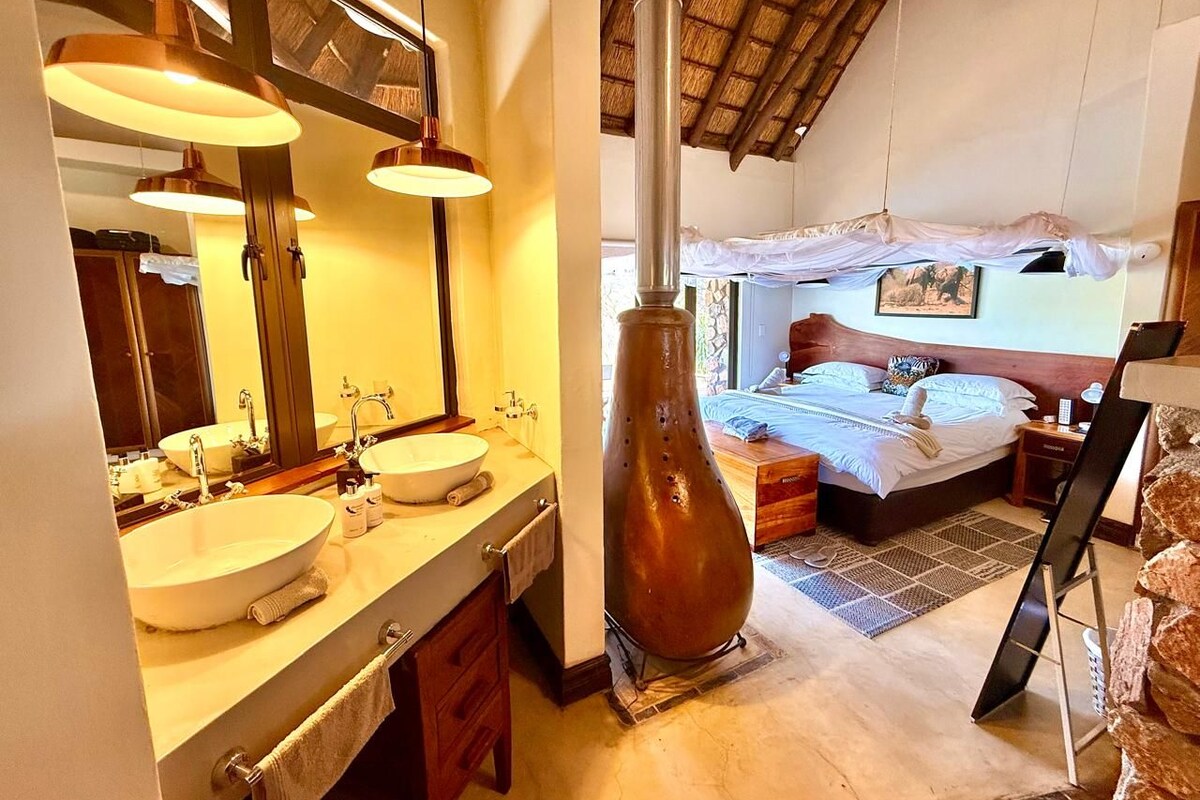 A bathroom with two sinks and a large mirror, a bedroom with a bed and a nightstand, and a unique decorative vase.