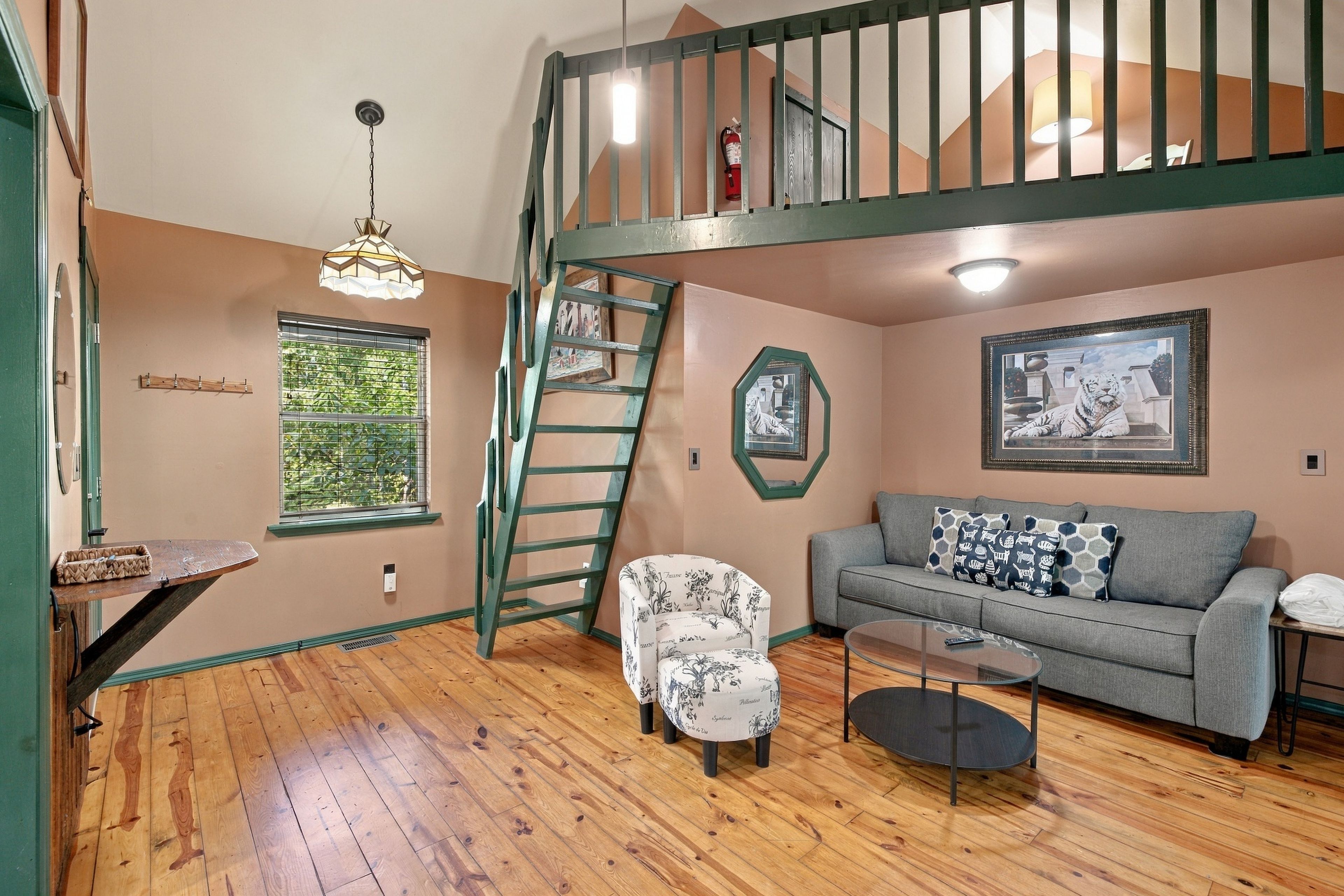 A room with a wooden floor, a grey sofa with patterned cushions, a white armchair, a glass coffee table, a ladder, a hanging light fixture, and a framed picture on the wall.