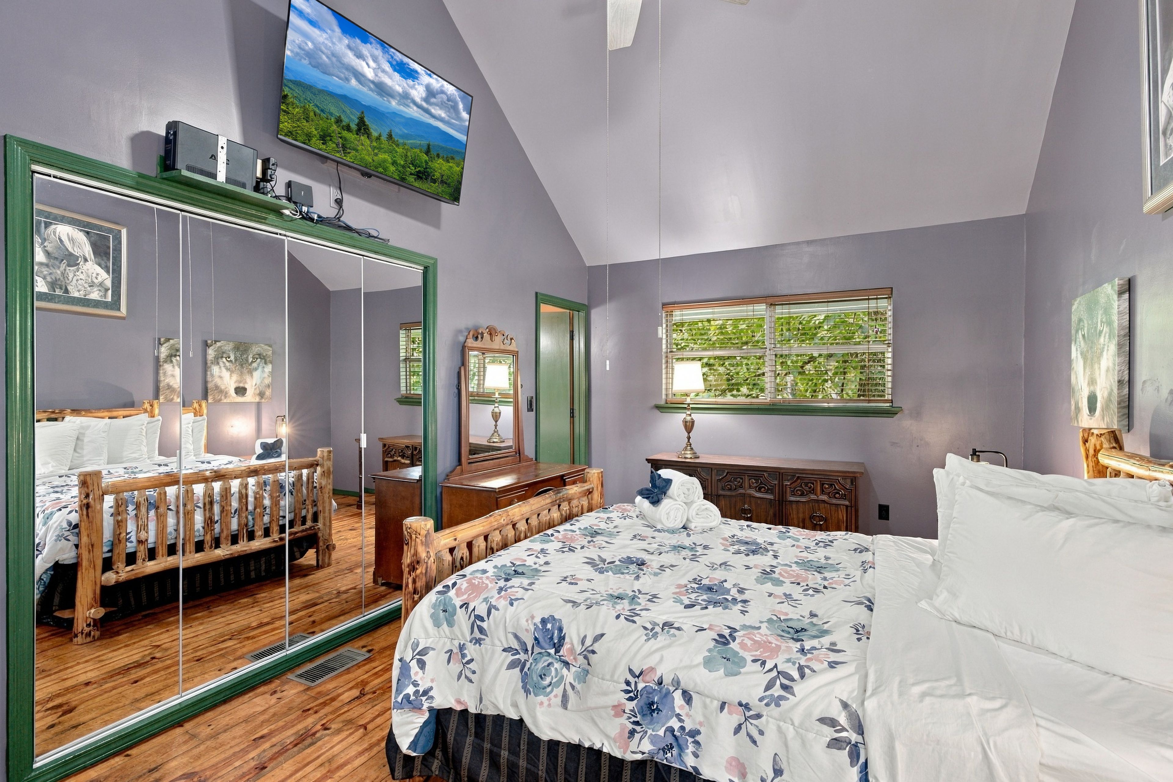 A bedroom with a large mirror, a bed with floral bedding, a dresser, and a window with a view of greenery.