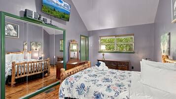 A bedroom with a large mirror, a bed with floral bedding, a dresser, and a window with a view of greenery.