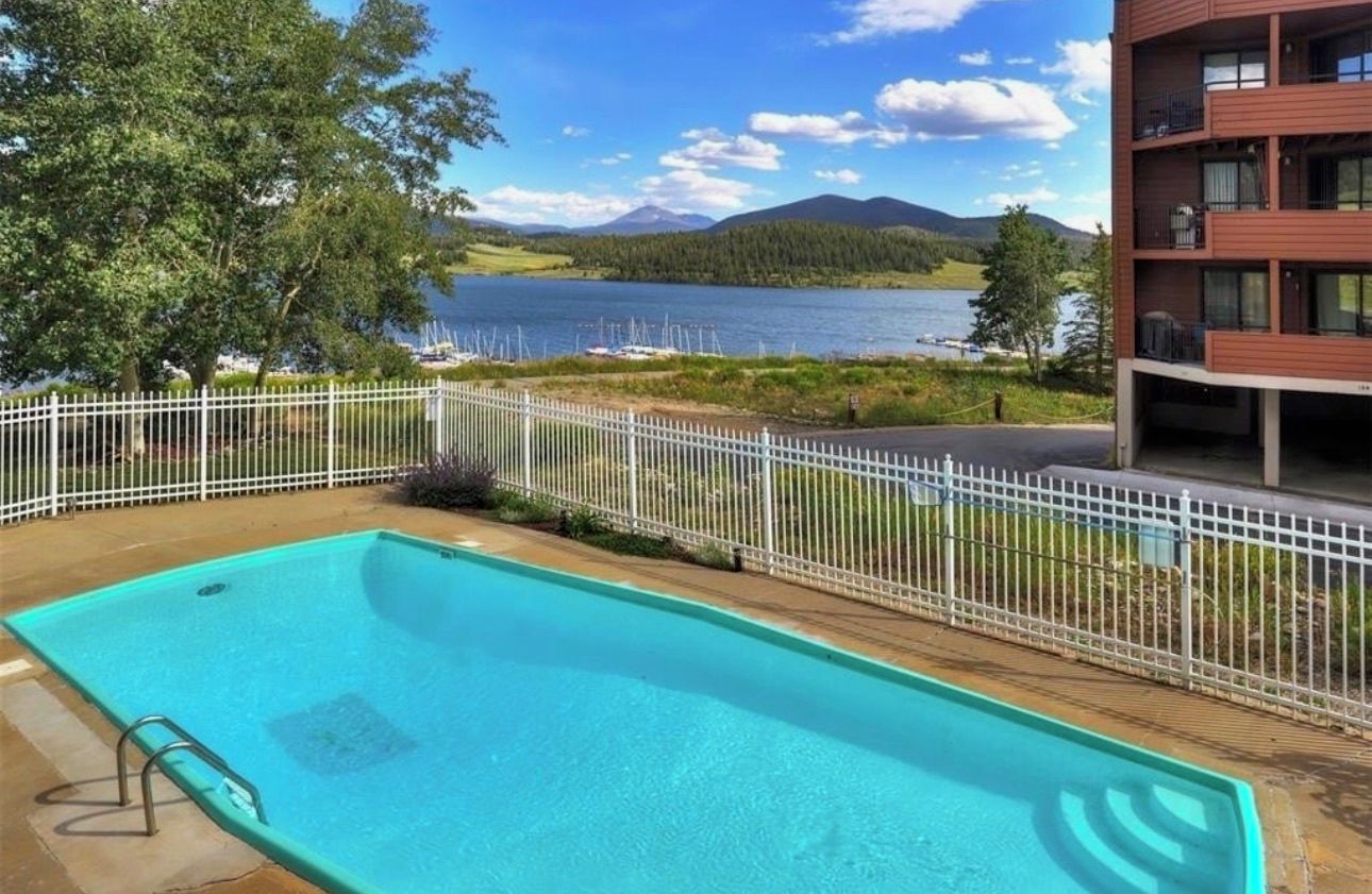 Una piscina con cerca blanca, un edificio con balcones y vistas a un lago y montañas.