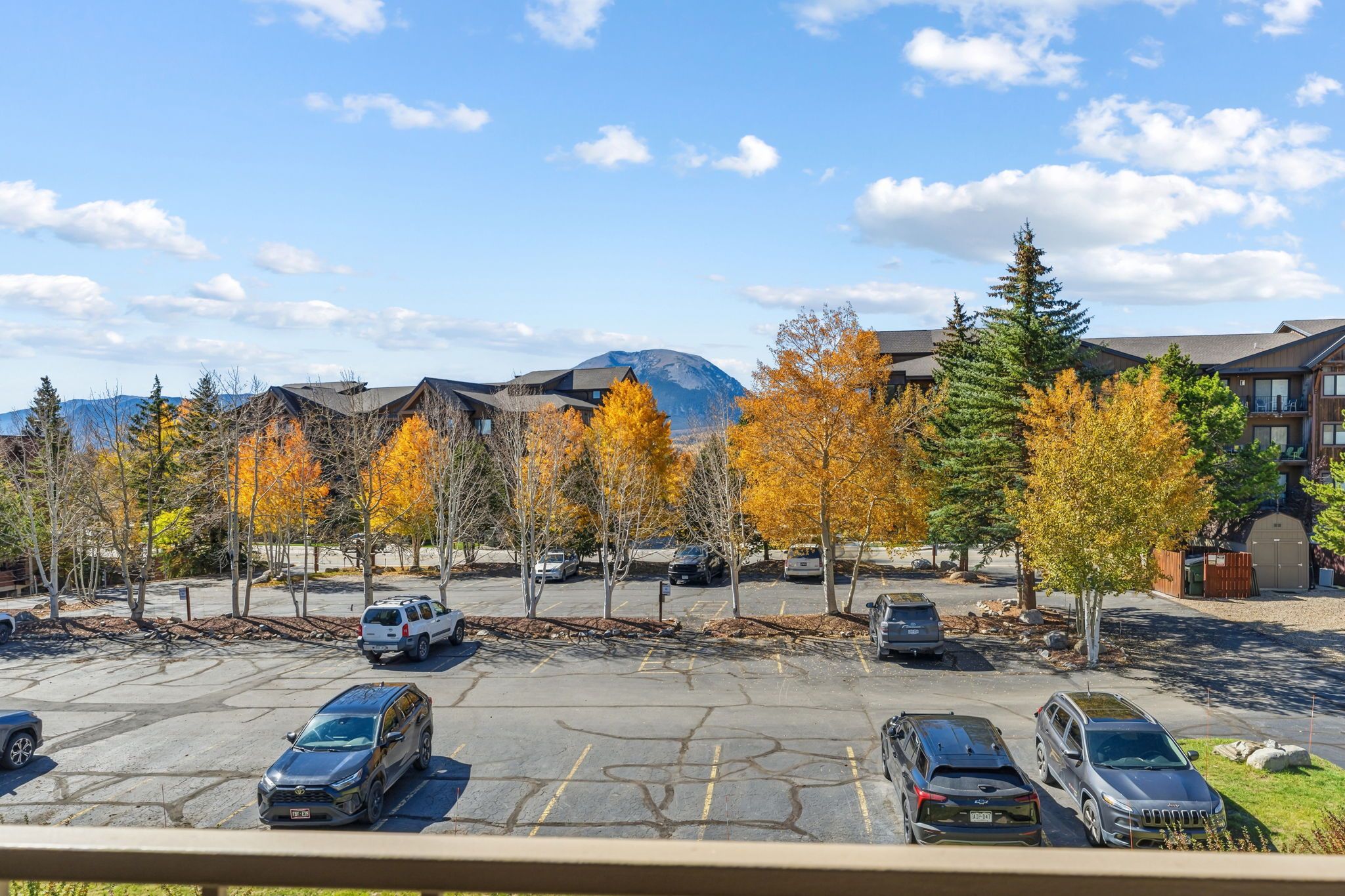 Ein Parkplatz mit mehreren Autos, umgeben von Bäumen mit Herbstlaub, und Gebäuden im Hintergrund.