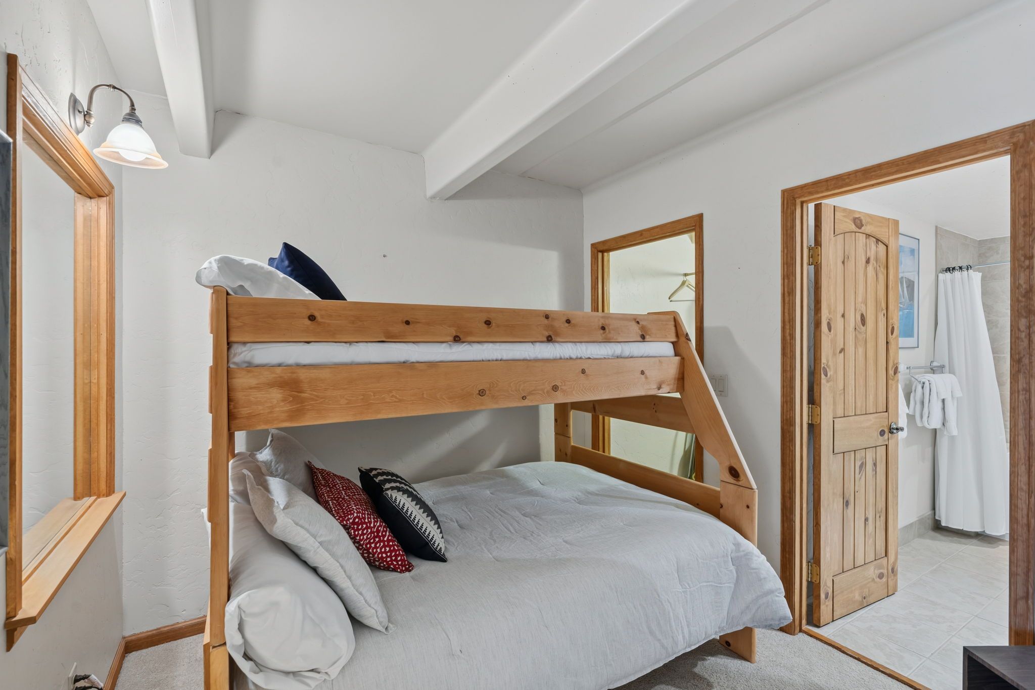 Una cama individual con estructura de madera, ropa de cama blanca y varias almohadas. Hay un espejo en la pared, una puerta de madera que da al baño y una lámpara sobre la cama.