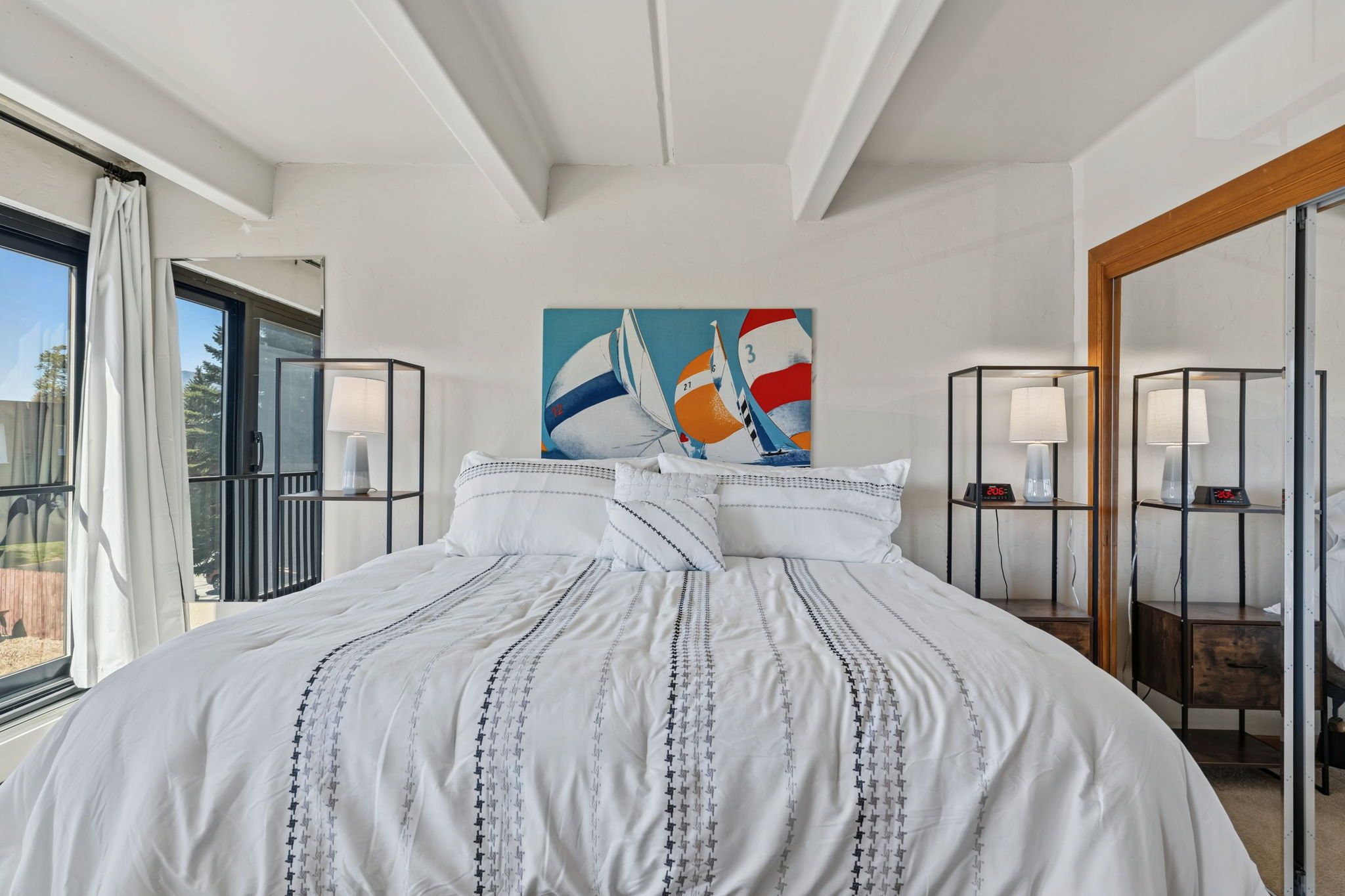A hotel room with a large bed, a painting of sailboats, a window with curtains, and a nightstand with a lamp.