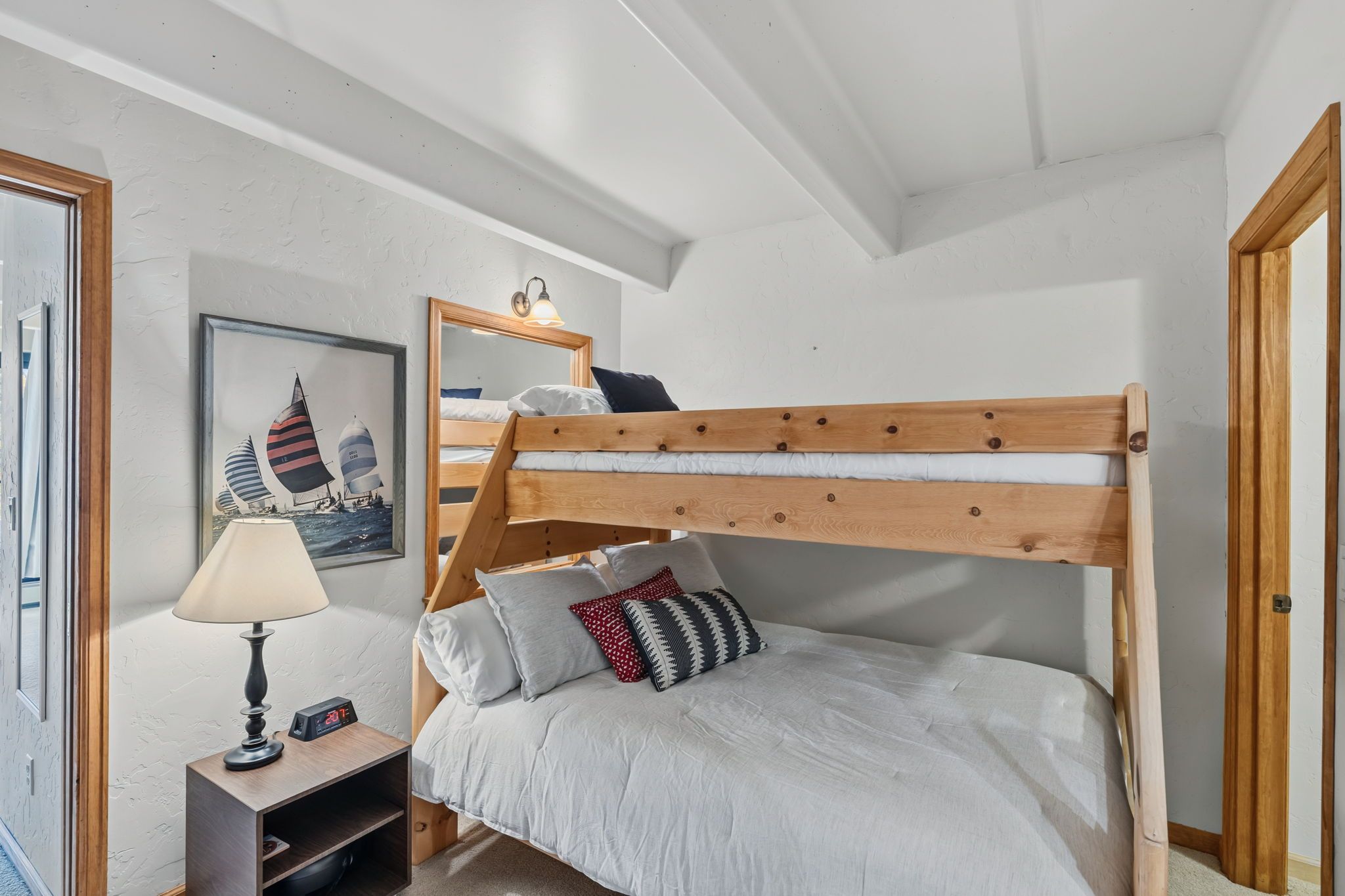 Una cama litera con estructura de madera y ropa blanca, una mesita de noche con lámpara, y un cuadro enmarcado en la pared.