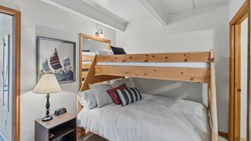 Una cama litera con estructura de madera y ropa blanca, una mesita de noche con lámpara, y un cuadro enmarcado en la pared.