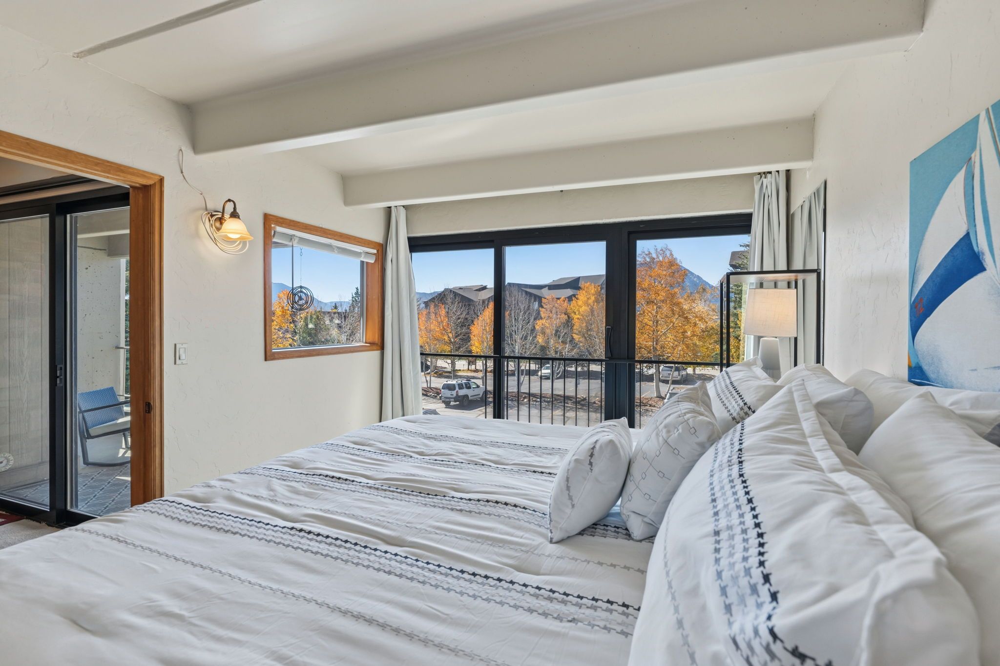 Habitación de hotel con una cama grande, un balcón con vistas a árboles otoñales y un cuadro en la pared.