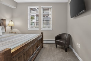 A bedroom with a bed, a chair, a TV mounted on the wall, and two windows with blinds. - Premium Unit 3301 - Two Bedroom - Fraser Crossing/Founders Pointe 2 Condo (Winter Park)