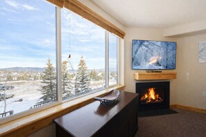A cozy living room with a fireplace, a large window with a snowy view, and a flat-screen TV mounted above the fireplace. - Heart Of Canyons Village Walk To Lifts 226 1 Bedroom Condo (Park City)