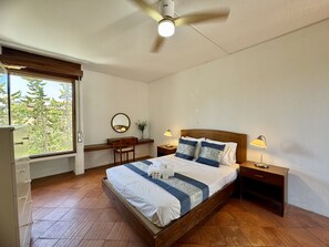 Ein Schlafzimmer mit einem Bett, Nachttischen, einem Schreibtisch, einem Stuhl, einem Deckenventilator und einem Fenster mit Blick auf Bäume.